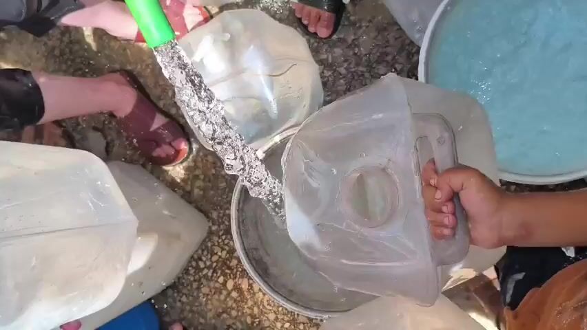 A group of people are gathered around filling large clear plastic jugs with water from a hose. The water is being poured into several jugs, some of which are already partially full. The people are using their hands and holding the jugs to position them under the stream of water. Some of the jugs have green spouts. The ground appears to be gravel or dirt. People's feet and legs are visible, wearing sandals and other casual footwear. The scene suggests a communal effort to collect water.