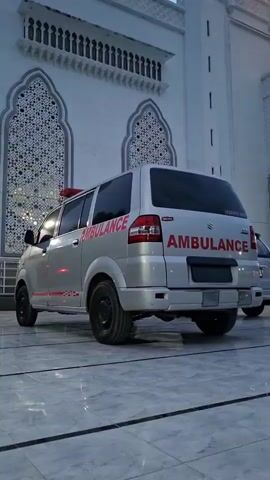 The video shows the rear side view of a silver ambulance parked on a paved surface. The ambulance has "AMBULANCE" written in red lettering on its side and rear. A red flashing light is visible on the roof of the vehicle. The background features a white building with decorative arched windows and a darker, textured ceiling or overhang. The overall lighting suggests it is either dawn or dusk. There are no people visible in the frame.
