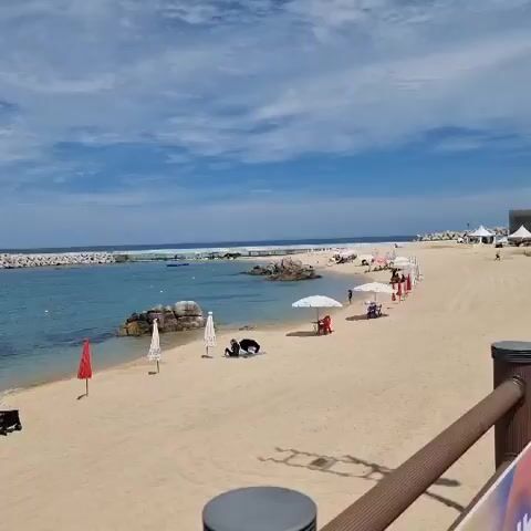 The video shows a beach with sand, ocean, and sky. There are several beach umbrellas, some of which are red. There are a few people on the beach, some are sitting under umbrellas, and one person is walking near the water. There are also some rocks and a lighthouse in the distance. The sky is blue with white clouds. The water is clear and blue.