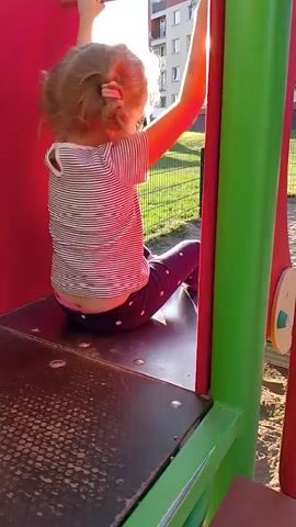A young girl with blonde curly hair is sitting on a platform of a playground structure. She is wearing a striped t-shirt and dark leggings with polka dots. She is looking at the camera and holding onto the railing of the structure with her hands. The playground structure is red and green. In the background, there is a building, grass, and a fence. The sun is shining.