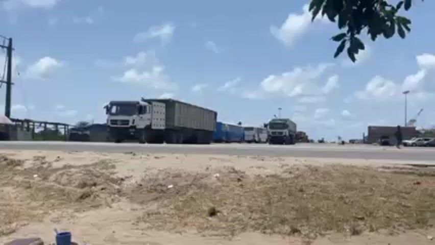The video shows a road with a line of trucks and buses. The sky is partly cloudy and the sun is shining. In the foreground is a dry, grassy area. A silver station wagon passes by the camera. To the right of the road are several shipping containers and port cranes. There is a person standing by a car near the containers. A small, blue cup sits on the ground in the very foreground. The video is stable but shaky. The leaves of a tree obscure the upper right-hand corner of the frame.