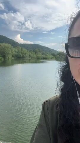 A video shows a partial view of a person on the right side of the frame, capturing a scenic landscape. The person has long, dark, curly hair and is wearing sunglasses and a dark green top.

The main focus of the video is a wide, calm body of water, likely a river or lake, stretching across the middle ground. Its surface shows subtle ripples, reflecting the bright sky. Beyond the water, lush green hills covered with trees rise into the distance under a blue sky dotted with scattered white clouds. The overall impression is one of a serene natural environment.

The video appears to be taken during daylight hours, likely in the late morning or afternoon, given the bright light. The weather seems pleasant and clear. The person is static, seemingly observing the view.

Based on the provided context, the location is Kamianytsia, Ukraine. No text is visible in the video.