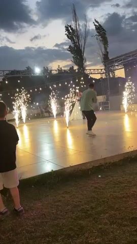 This video captures a wedding reception outdoors at dusk. The focal point is a couple, presumably the bride and groom, dancing on a illuminated stage. The bride wears a white wedding dress and veil, and the groom a dark suit. Surrounding them, several pyrotechnic fountains are erupting, creating a dramatic and celebratory atmosphere. In the foreground, a young boy in a black shirt and white shorts is visible, with his back to the camera, appearing to be playing or watching the event. In the background, string lights are strung across the venue, and tables and chairs suggest other guests are present. The sky is overcast with clouds, and trees are visible behind the stage. The overall scene is festive and romantic, set against the backdrop of twilight.