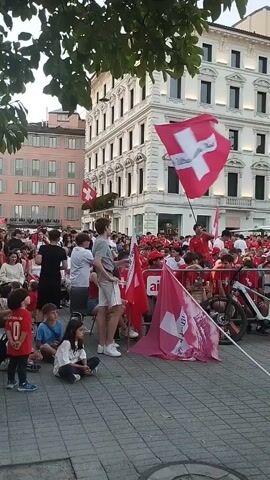 A group of people are gathered outdoors, many wearing red shirts, and some holding Swiss flags. In the foreground, a young person in a gray t-shirt and shorts holds a flag, while other children sit on the ground. A white bicycle is parked to the right, and a metal barrier separates the crowd from the street. In the background, buildings with white and beige facades line a street, and trees provide shade. The scene appears to be a gathering or celebration.