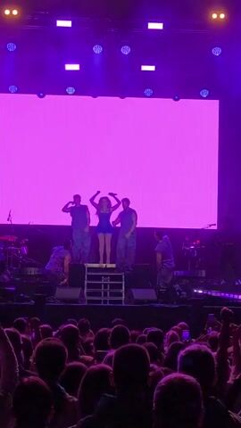 A female vocalist performs onstage with several backup dancers and musicians, with a crowd of people watching the performance. The vocalist stands on a platform towards the center of the stage. She wears a short blue dress and holds a microphone. She has her arms raised. Behind her, a large screen shows a purple background with bright white circles appearing. To either side of the singer are three male backup dancers in t-shirts and pants. To the left, a drummer plays. The stage is lit with purple lights and some white lights overhead. The crowd of people in the foreground are mostly dark silhouettes due to the bright stage lights. Some have their arms raised.