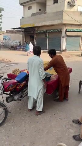 The video shows a street scene in a sunny area, possibly in a village. A man in a light-colored shalwar kameez stands near a red cart attached to a red motorcycle. The cart is piled with a heap of what appears to be yellow granular material. Another man, dressed in a brown shalwar kameez, is using a hand scoop to transfer the yellow material from the cart into a red bag. A small, blue mechanical scale sits on the cart's surface. In the background, people walk along the street, and buildings with closed metal shutters line the street. Another man is seen walking in the background, carrying a large sack over his shoulder. The road surface is dusty and shows signs of wear.