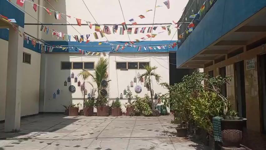 The video shows a courtyard area of a school building. The courtyard is paved with tiles and has a few potted plants and trees. The walls of the building are light yellow with blue trim. A line of small flags from various countries is strung across the courtyard overhead. On the walls, there are some decorative items, possibly clocks or dartboards, and a few posters. There are trash cans visible in the foreground. In one section of the building, there are several doors and windows, and a shoe rack is placed outside one of the doors. In the background, there are a few people sitting and walking. The lighting suggests it is daytime, and the scene appears to be outdoors.