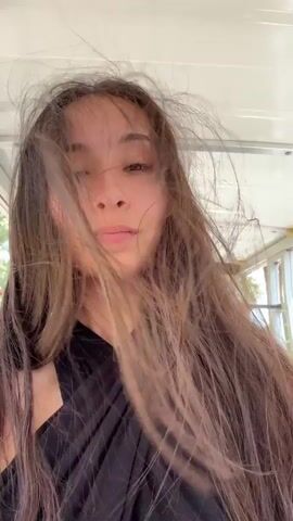 The video shows a young woman with long brown hair blowing in the wind. She is wearing a black top. She is in the passenger seat of a vehicle, possibly a golf cart, as indicated by the white roof and metal railing visible in the background. Another person, a male, is visible in the driver's seat, wearing a white baseball cap with the number 95 on it and a dark t-shirt. The environment appears to be outdoors, with some greenery and buildings visible through the windows of the vehicle. The woman is looking towards the camera and smiling slightly. The footage is shaky, suggesting the vehicle is in motion.