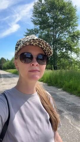 A woman with long blonde hair, wearing a grey t-shirt, sunglasses, and a leopard-print cap, stands on a paved path. She is looking at the camera and smiling. To her left is a grassy area and trees. To her right is a wider paved path with trees and a blue sky with some clouds. The camera moves, showing more of the path and trees.