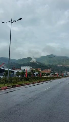 The video shows a wide shot of a road on a cloudy day in Vietnam, with a large white Buddha statue situated on a hill in the background. The road in the foreground is wet, presumably from rain, and lined with green bushes. Two tall streetlights flank the left side of the road. Several buildings are visible between the road and the base of the hill. Red flags are planted near the road. The Buddha statue, the primary focal point, is surrounded by the green foliage of the hill and partially obscured by the low-hanging clouds. The scene is peaceful, with a focus on the contrast between the urban elements and the natural landscape featuring the prominent spiritual figure.