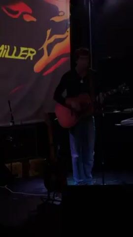 A young man with dark curly hair is performing on stage at a music venue. He wears a black long-sleeved shirt and blue jeans, and he plays an acoustic guitar. He stands center stage, illuminated by stage lighting, singing into a microphone. Behind him is a screen with abstract, colorful imagery projected onto it. To his left, slightly behind him, is another guitar on a stand. A keyboard and other equipment are visible to his right. The room is dimly lit, suggesting a concert or performance setting.