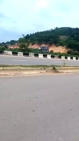 The video shows a highway scene. The camera is positioned on the side of the road, facing the highway with a green, tree-covered hill as a backdrop. The road is asphalt and has a white line painted on it. There's a black and white striped barrier between the road and the hill. At one point, a person walks along the side of the highway near the barrier. The sky is overcast. There is significant wind noise throughout the video.