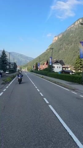 A motorcycle is riding down a road. The motorcycle is blue and black and has a rider wearing a helmet. The road is asphalt and has white lines marking the lanes. There are houses and trees on the side of the road. In the background, there is a large mountain covered in trees. The sky is blue and there are some clouds. There are also some banners hanging from poles along the side of the road. A pedestrian crossing is visible. The scene appears to be in a rural or semi-rural area.