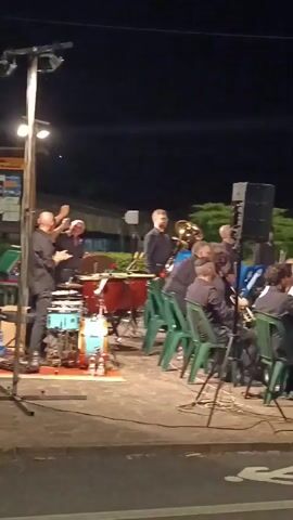 The video shows an outdoor evening performance by a band. The band members, all wearing black shirts, are seated on green chairs arranged in rows. They are playing various instruments, including drums, brass instruments like trumpets and trombones, and possibly a saxophone. In the foreground, there are speakers and a projector placed on top of a green trunk. The background is dark, indicating it's nighttime, with some trees and foliage visible. The ground appears to be asphalt, with a white pedestrian crossing symbol painted on it. The band members appear to be engaged in their performance, with some looking at sheet music. The overall atmosphere suggests a public event or concert.