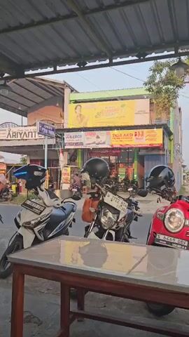The video shows an outdoor scene with three motorcycles parked in front of a building. The motorcycles are a white scooter on the left, a black and white motorcycle in the middle, and a red scooter on the right. All three motorcycles have helmets on their handlebars. The building behind the motorcycles has a yellow banner across the top with text in Indonesian. There are people and other motorcycles in the background, suggesting a busy area. The lighting suggests it is daytime, possibly late afternoon. A table with a tiled surface is in the foreground, obscuring the lower part of the motorcycles. The scene appears to be a street-side establishment.