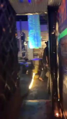 The camera pans left from a dimly lit open doorway to reveal a courtyard-style outdoor cafe or lounge. The space has a bohemian vibe, with low seating made from tires and pillows, and potted plants scattered around. A translucent, shimmering decoration hangs from the roof, illuminated with blue and green lights. There's a building in the background with multiple windows adorned with colored glass. The overall atmosphere is relaxed and inviting.