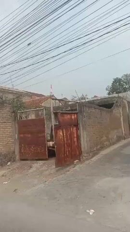 The video shows a first-person view from a motorcycle traveling down a street lined with houses and trees. Several Indonesian flags are displayed, suggesting a national holiday or celebration. There are other vehicles on the road, including a car and another motorcycle. The road is paved, and there are utility wires overhead. The weather appears to be overcast. The video ends as the motorcycle approaches an archway decorated with flags and banners.