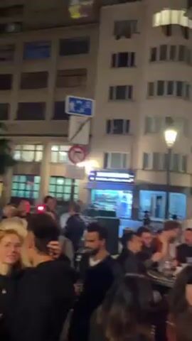 The video shows a crowded scene outside a bar called "Le Belgica" at night. Many people are gathered, standing and talking in small groups. Some are holding drinks. The bar has a red awning and is well-lit. There are buildings visible in the background and a streetlamp. The camera moves and captures different angles of the crowd and the bar's facade.