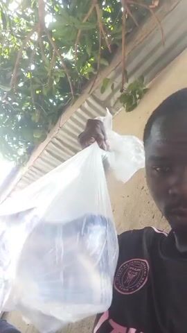 A man is holding a plastic bag of ice in his right hand. He is wearing a black Adidas shirt with pink stripes. He appears sweaty. The background shows a tree, a corrugated metal roof, and a concrete wall.