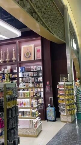 The video pans across a brightly lit duty-free shop, showcasing a variety of perfumes, cosmetics, and souvenir items. The shelves are meticulously organized, with products displayed in an aesthetically pleasing manner. In the first few seconds, the camera focuses on elaborate perfume bottles and ornate gift boxes. A man in a suit and hat walks through the shop, browsing the merchandise. The scene then shifts to a wider view of the airport terminal, with people walking by and signs indicating lounge areas. The architecture of the terminal is modern and spacious, with high ceilings and sleek design elements. The overall impression is one of a high-end retail environment within a busy international airport.
