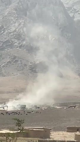 A dust devil swirls in front of a mountainous background. In the foreground, a small collection of buildings lines a road. There are some animals in front of a machinery in the dust.