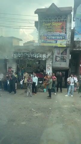 The video depicts a street scene in Kangra, India. Pedestrians, including adults and a child, are standing or walking along the roadside near storefronts and signs advertising businesses like "Swar Sagar Music Academy" and "Fortis Hospital Kangra." Stalls selling produce are set up along the side of the road. The road is lined with parked cars and vehicles traveling through the area. The sky is overcast, and the sunlight is bright. The video appears to be taken from a moving vehicle.