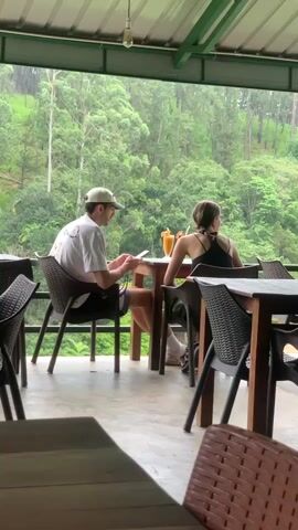 A man and a woman are seated at tables on a covered outdoor patio, looking out at a lush, green, and misty forest-covered mountainside. The man, wearing a white t-shirt, black shorts, and a light-colored baseball cap, is seated on the left. He is looking down at his phone, which he holds in his hands. A glass with a yellow drink and a straw is on the table in front of him.

The woman, wearing a black tank top and dark shorts, is seated at the table to the right of the man. Her back is to the camera, and she is looking out at the view. A glass with a yellow drink and a straw is on the table in front of her. The tables are made of wood, and the chairs are made of dark-colored wicker.

The patio is covered by a metal roof with a green trim, supported by wooden posts. The floor of the patio is a light-colored concrete. The scene appears to be set in a restaurant or cafe overlooking a scenic natural landscape. The misty, overcast weather and the dense green foliage suggest a tropical or subtropical environment, consistent with the location context of Ella, Sri Lanka. The overall mood is peaceful and relaxed. There is no visible text in the image.
