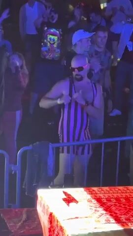 A bald man with a beard, wearing a pink and black striped singlet and sunglasses, dances in front of a crowd at an outdoor event. The crowd is visible behind a metal barrier. The man is smiling and gesturing with his hands. In the foreground, there is a table covered with a red patterned cloth, with a few small white objects on it. The lighting suggests it is nighttime.