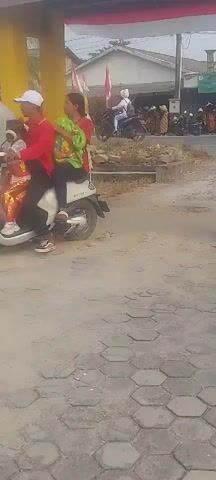 This is a short video showing a street scene, likely in a village or small town. The foreground is a dirt and paved road with hexagonal pavers. In the mid-ground, several people are visible on and around motorcycles. There are red and white flags displayed on poles, suggesting a local event or celebration. A yellow building structure is on the left.

At 00:00, a motorcycle with two people on it is seen from behind. The person in front is wearing a white shirt and dark pants, and the person behind is wearing a green top and is partially obscured.

At 00:01, a motorcycle with a rider wearing a patterned shirt and helmet is seen from the side.

At 00:02, a motorcycle with a rider and a child passenger is seen. The rider is wearing a helmet and a red top, and the child is also wearing a helmet.

At 00:03, a motorcycle with three people on it is visible. The rider is wearing a helmet and red pants, and two children are on the motorcycle, one in front and one behind. The children appear to be wearing some type of uniform or official attire.

At 00:04, the scene shows people on motorcycles and standing near a fence. Two individuals are on a motorcycle with a rider in a helmet and red pants, and a child in front. Another person, wearing a red shirt and dark pants, is standing to the right of the yellow structure. Another man in a light-colored shirt and brown pants is also standing nearby. The environment appears to be an outdoor gathering or procession.