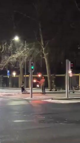 A man is juggling fire at night. He stands on a snowy sidewalk near a traffic light and juggles three torches with flames. Other traffic signals are visible nearby, and a car passes in the street. The scene is lit by streetlights and reflected in the wet pavement. Bare trees and building facades are in the background. A person sits on a scooter to the left of the man.