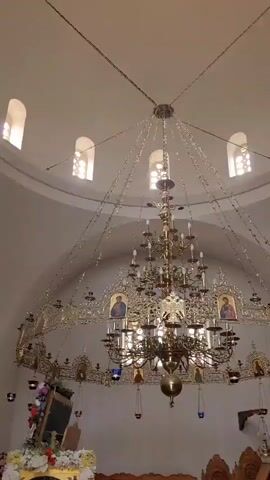 This media file captures the interior of an Orthodox church, likely in Monagrouli, Cyprus, given the context. The primary focus is a large, ornate chandelier hanging from the domed ceiling. The chandelier is made of brass and features numerous candleholders, some of which are lit. It is adorned with icons of religious figures.

The church interior is characterized by a domed ceiling with arched windows that admit light. The walls are painted a light color, contributing to the overall illumination. To the right of the chandelier, a richly carved iconostasis is visible, decorated with more religious icons and intricate woodworking. Smaller lamps and decorative elements are also present throughout the space.

The scene appears to be calm and reverent, suggesting a quiet moment within the church, possibly outside of a service. The lighting suggests it could be daytime, with the natural light from the windows complementing the artificial light from the chandelier and other lamps. The overall impression is one of solemn beauty and spiritual significance, typical of Orthodox church interiors. There is no visible text in the media file.