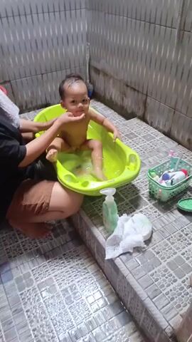 A person is bathing a baby in a green plastic tub. The baby is sitting in the tub, which contains water. The person is holding the baby's legs. There are toiletries and a diaper next to the tub. The scene appears to be a bathroom, with tiled walls and floor.