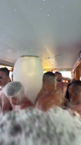 This video captures a vibrant foam party taking place on the interior deck of a boat in Menderes, Turkey, during the daytime. Numerous young to middle-aged adults, predominantly men, are visible, many shirtless or in swimwear, completely covered in white foam. A large foam machine in the center of the boat continuously generates more foam, enveloping the attendees.

Participants are seen laughing, smiling, and dancing amidst the foam, appearing to enjoy the festive atmosphere. The boat's interior features wooden paneling and windows offering glimpses of the clear blue sea and sky, confirming the maritime setting. In the background, some individuals are observing, while others are seen with drinks and a hookah on a table. Partial text on a sign in the background reads "BATHROOM BOAT TOUR," "BATHROOM," and "SUGGESTED," suggesting the type of excursion. Upbeat music plays throughout the scene, enhancing the party mood.