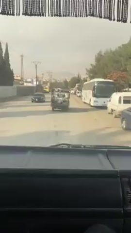 From inside a vehicle, likely a car or truck given the dashboard view, the video shows a street scene in what appears to be a Middle Eastern or Mediterranean setting.  There is traffic, including a white tour bus, a dark sedan, and a small truck with some sort of cargo. Power lines are visible, and there are green trees on the sides of the road.  A distant hill or mountain is visible under a hazy sky.  A person's leg is briefly visible in the lower right of the frame.