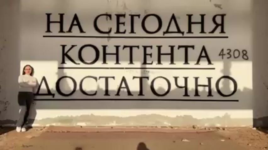 A woman stands in front of a wall with Russian text painted on it. The text reads "НА СЕГОДНЯ КОНТЕНТА 4308 ДОСТАТОЧНО." The woman is wearing glasses, a white long-sleeved shirt, and dark pants. She is leaning against the wall with her arms crossed, then puts her hands behind her head. The wall is white with some damage at the bottom. The ground in front of the wall is a mix of dirt and debris. Sunlight is casting shadows across the wall.