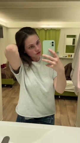 A young woman with light skin, wearing a light gray t-shirt and dark jeans, stands in a bathroom and takes a selfie with her light green Apple iPhone. She runs her right hand through her dark brown hair, which is partially pulled back.  The bathroom features light-colored wooden floors, a white sink, a large mirror reflecting the room, and green curtains. A bed and a small wall shelf are also visible in the reflection. The lighting in the room is muted and even.