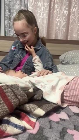 A teenage girl is playing with a baby on a bed. The girl is wearing glasses, a denim jacket, and has her hair in a braid. She is sitting up in bed, looking down at the baby. The baby is lying on its back, covered with a blanket. The baby is wearing a patterned pajama set. The girl is holding the baby's hand and talking to her. The bed is covered with a patterned blanket and there are pillows behind the girl. The room appears to be a bedroom with curtains on the window.