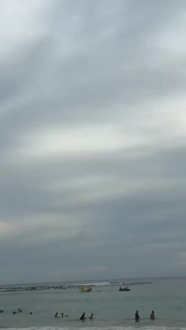A wide shot shows a beach with gray clouds in the sky. In the shallow water near the shore, several people are wading. Further out in the ocean, a yellow inflatable object shaped like a cartoon character is being towed by a jet ski. Another jet ski is nearby. The water is a light turquoise color and there are small waves breaking on the shore.