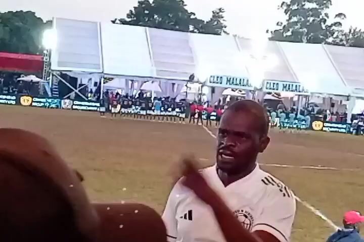 The video depicts an outdoor scene, possibly a sporting event. In the foreground, a man wearing glasses and a white Adidas shirt looks towards the camera. Behind him, there's a grassy field with two rows of people standing in the distance, possibly teams lined up. White tents are set up, with signage that reads "CLEO MALALA" visible on them. Bright lights illuminate the scene. The sky is overcast. The camera appears to be held by someone standing in the crowd.