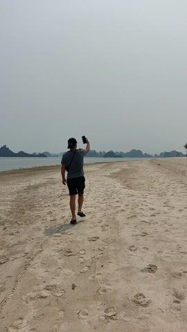 The video shows a man walking on a beach, away from the camera, holding a mobile phone. He is wearing a grey t-shirt, black shorts, a black baseball cap, and black sandals. He also has a black bag slung across his shoulder. The beach is sandy with many footprints. In the background, there is a body of water with several small islands or rock formations. The sky is overcast.
