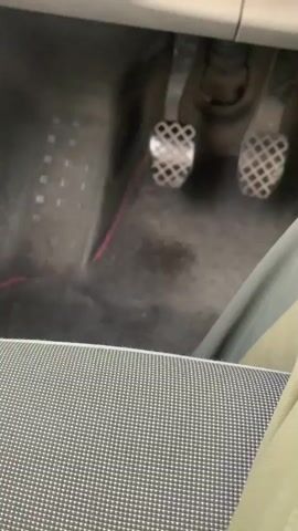 A blurry video shows the interior of a car from the perspective of someone sitting in the passenger seat. The camera is focused on the floor area in front of the driver's seat, specifically on the pedals. There are two metal pedals visible, one on the left and one on the right, both with a diamond-patterned grip. A thin red wire is visible running across the floor. The floor itself appears dusty and somewhat dirty. In the foreground, the edge of a patterned car seat is visible. The lighting is dim. No people are visible in the frame.