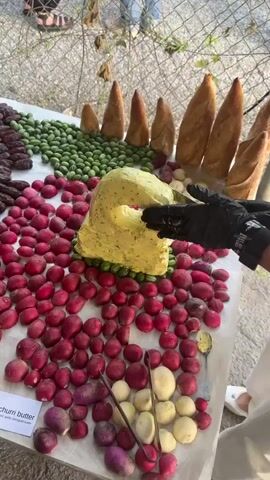 The video shows a table laden with food items, arranged in a decorative manner. In the foreground, a person wearing black gloves is using tongs to pick up food from the table. The table itself is covered with a white cloth, and behind it, a wire fence is visible.

The food display includes several mounds of yellow, speckled butter, shaped into abstract forms. One large mound is surrounded by red radishes, while another, smaller mound sits atop a bed of dark brown dates. To the left of the dates, there is a dense arrangement of small, green olives, and further to the left, a row of dark, elongated objects, possibly more dates. Behind the green olives, several large baguettes are propped up, resembling a fence or a decorative element.

In the center of the table, a large quantity of radishes are arranged in a semi-circle. To the right of the radishes, there are several small, light-colored potatoes, some of which appear to be cut in half. Further to the right, more baguettes are visible.

Small white labels are placed on the table, one of which reads "Chimichurri Butter". The ground surface appears to be gravelly. Two people are partially visible in the background, standing behind the table and fence. The overall impression is that of a food tasting or presentation.
