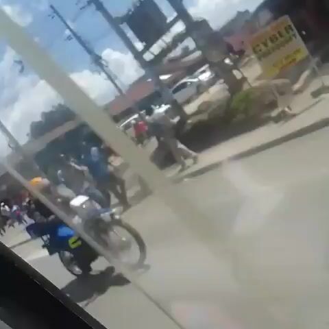 The video shows a view from inside a moving vehicle, likely a car, looking out onto a street scene. The camera angle is slightly tilted, and there are occasional reflections on the window. The video starts by showing a street lined with shops and pedestrians. There are several motorcycles parked or moving on the street, some with riders wearing helmets and delivery boxes. A few cars and vans drive by. The buildings have a mix of architectural styles, and there is a shop named "Mugwan Stores" visible. There are people walking on the sidewalks and some standing near the shops. There is a clear blue sky with some scattered clouds. As the vehicle continues to move, the view shifts to different parts of the street, showing more buildings, shops, and parked vehicles. The video ends with a view of a building with "PHARMACY" and "OPTICAL SHOP" signs.
