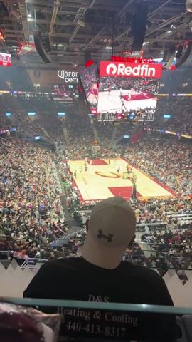 A high-angle shot displays a basketball game in progress at an indoor stadium. The basketball court is red and tan, with players in uniforms actively playing. The camera is positioned behind a spectator wearing a black shirt and a white Under Armour baseball cap. The stadium is filled with a large crowd of people.  Electronic scoreboards and advertisements, including "Redfin," are visible around the arena.