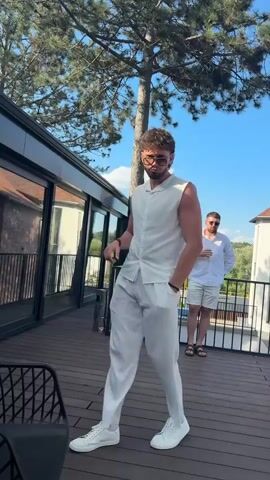 Two young men are on a wooden deck outdoors. The man in the foreground is wearing a white sleeveless button-down shirt and white pants, and white sneakers. He is facing the camera with his right arm raised and thumb up. He is also wearing sunglasses. In the background, another young man in a white shirt and shorts is visible, holding a drink. There is a chair in the lower left corner of the frame and trees and a building are visible in the background. The sky is blue with some clouds.