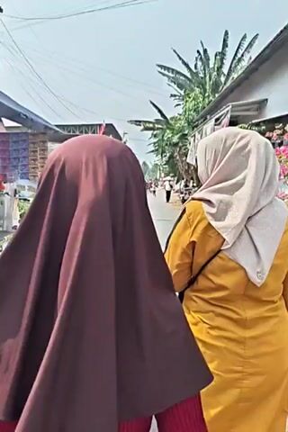 The video begins with a close-up of a street vendor's stall, displaying various packaged goods such as snacks and what appear to be personal hygiene products. The stall is covered by a blue tarp and positioned along a paved road. A person wearing a dark brown hijab is in the foreground, obscuring their face and partially blocking the view of the stall. To the right, another person in a yellow jacket and light-colored hijab is visible from behind, walking away from the camera. The road continues into the distance, lined with houses and trees. In the background, there are more people walking, some flags, and a large building with a metal roof. The sky is bright and appears to be daytime, with power lines visible overhead. The scene suggests an outdoor market or a community gathering along a street. The second half of the video shifts focus to a row of houses on the right side of the street, adorned with potted plants and flowering bushes, creating a more residential and tranquil atmosphere compared to the initial market scene.