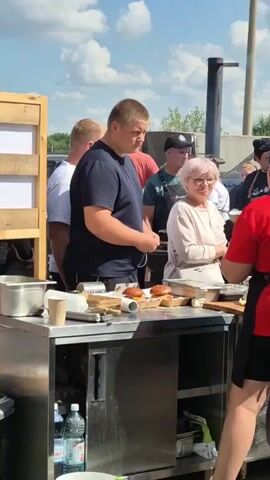 A group of people are gathered around a food preparation station outdoors. In the foreground, a young man in a dark blue t-shirt stands to the left, facing right. Beside him, a woman with short pink hair and glasses, wearing a light-colored cardigan, stands facing the young man. Behind them, several other individuals are visible, some wearing aprons, suggesting they are involved in food service or preparation. The station itself appears to be made of stainless steel, with various food items and containers on its surface, including what look like burger buns and condiments. In the background, there is a blue sky with white clouds, and some structures that suggest an outdoor event or market.