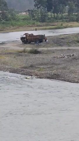 A dump truck is parked on the sandy bank of a river. Two people are standing near the truck, and one appears to be loading material into the truck bed. Another person is standing further away, closer to the water. The river is flowing, and there are trees and vegetation on the opposite bank. The weather appears overcast.