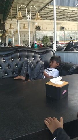 This media file shows an outdoor dining area, likely a restaurant, with many tables and chairs set up under a large, green-framed roof. The time of day appears to be daytime, with bright lighting evident. The scene is mostly empty, with only a few people visible. In the foreground, a young boy is asleep on a black leather sofa. He is wearing a white t-shirt and dark pants. Next to the sofa is a tissue box with a logo that appears to say "HACIENDA" in stylized text. In the background, there are rows of black tables and chairs, some of which are occupied by patrons. The setting is open-air, with a view of buildings and greenery beyond the dining area. The overall mood seems relaxed and casual, with the sleeping child indicating a peaceful moment within the busy environment. There is some visible text on signs in the background which is too blurry to read.