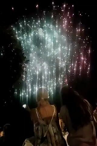 Two women are visible from behind, facing away from the camera, watching a fireworks display. The woman in the foreground has blonde hair and is wearing a floral dress. She is holding a phone and appears to be taking a photo or video. The woman next to her has long, dark hair and is wearing a light-colored shirt and pink pants. In the background, several fireworks are exploding in the night sky, creating streaks of green and pink light. The scene is dark, with the only light coming from the fireworks and the phone screen.